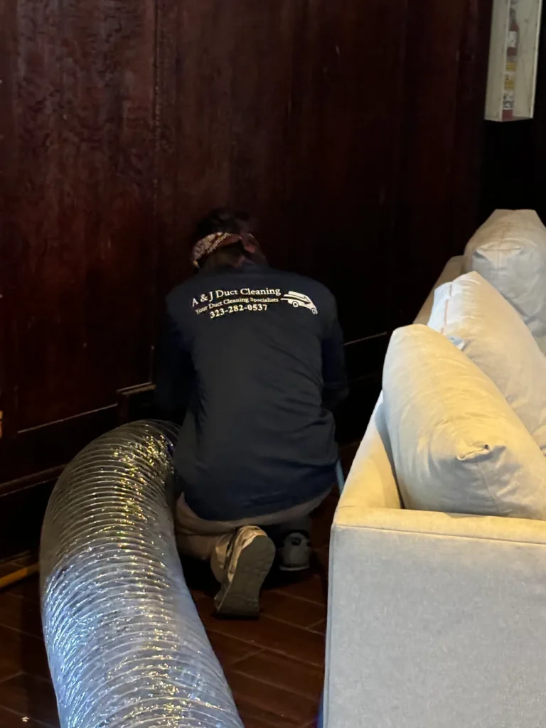 A technician kneeling behind a sofa to run the duct hose to a wall vent