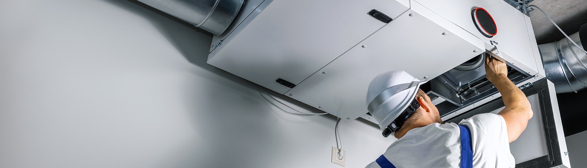 A technician on a ladder working on ceiling ductwork in a bright room
