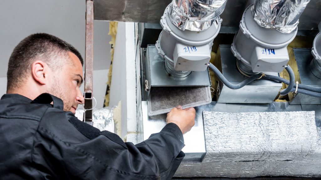 A technician in dark workwear removing a filter from an overhead duct unit fitted with flexible silver ducting