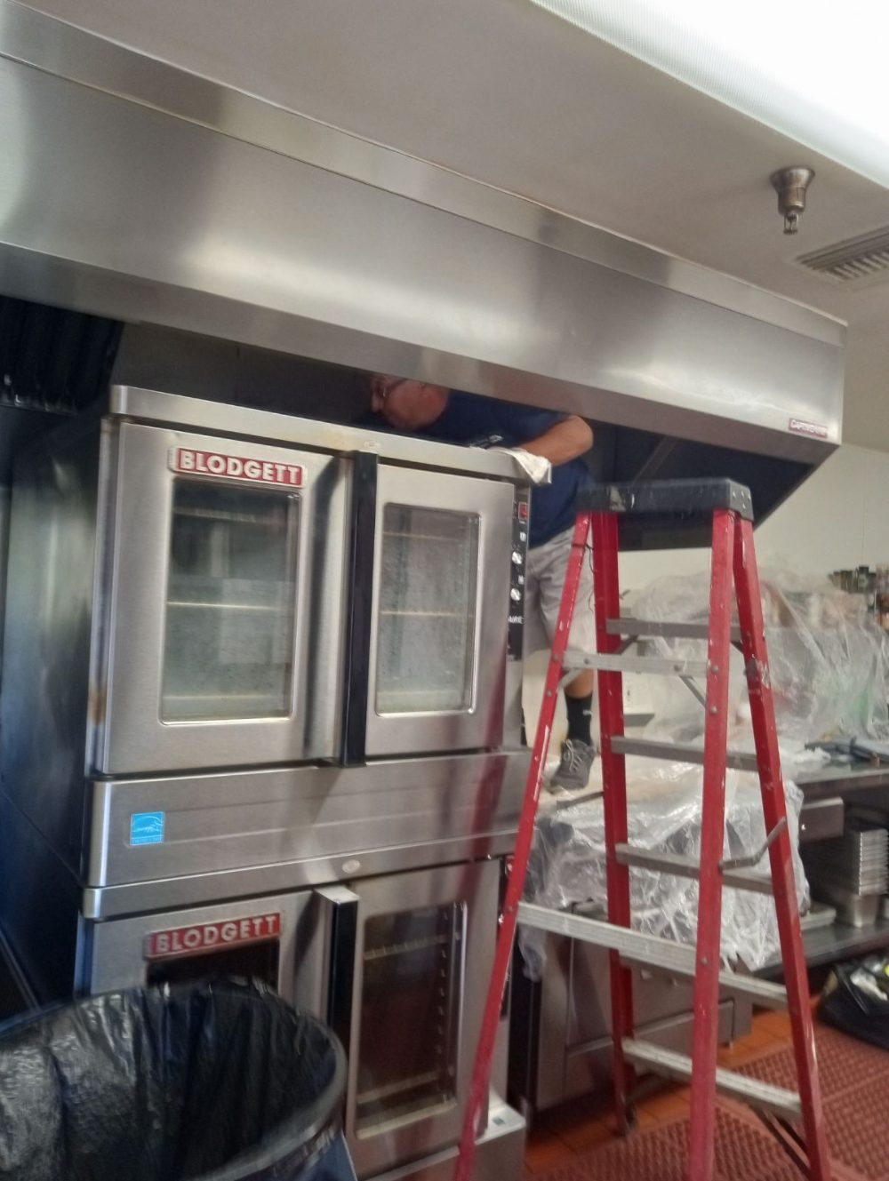 A technician on a step ladder cleaning a stainless kitchen exhaust hood above a commercial oven