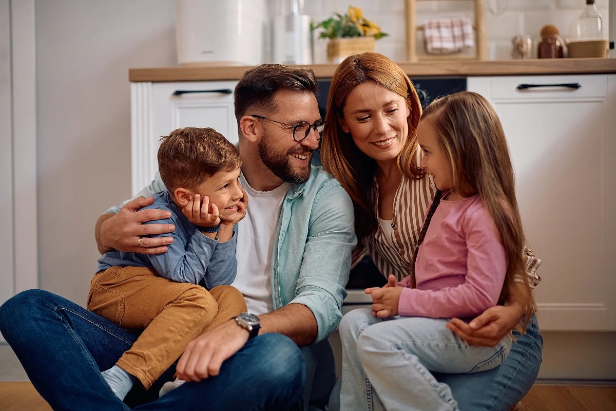 A family of four sitting together on the floor of a bright kitchen