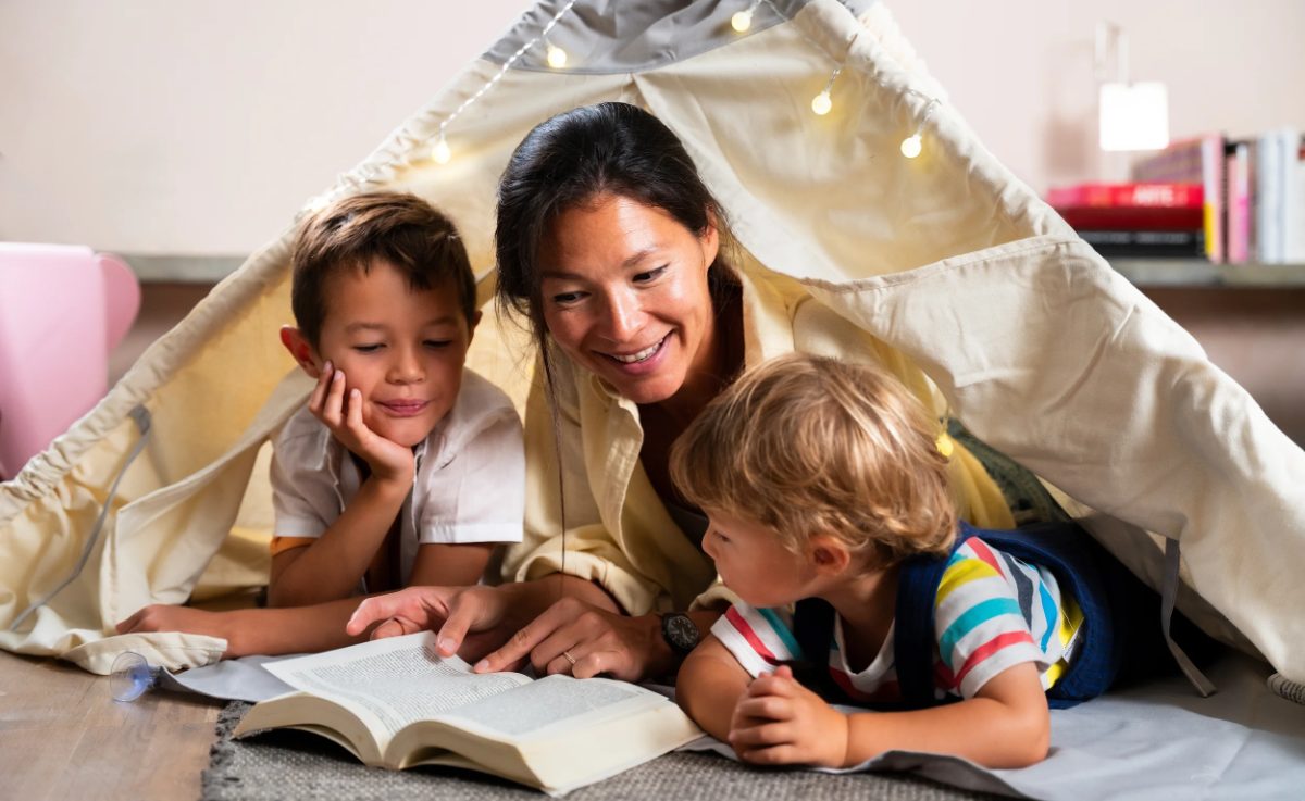 A parent and two children reading a book under a blanket fort