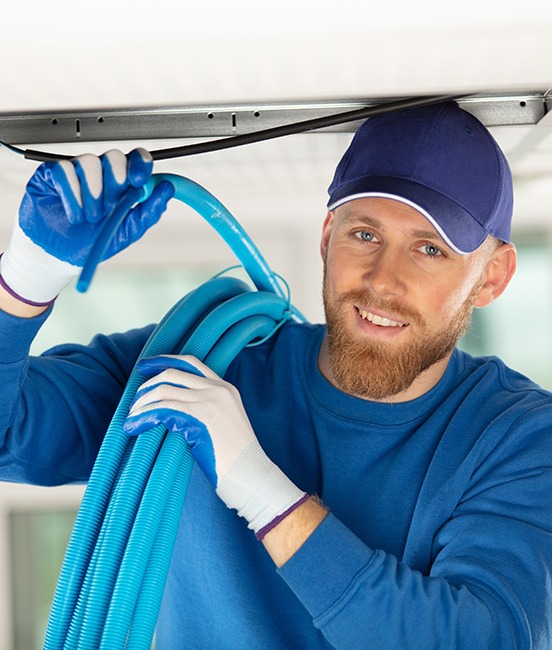 A technician in a blue cap carrying a duct cleaning hose beneath a ceiling vent