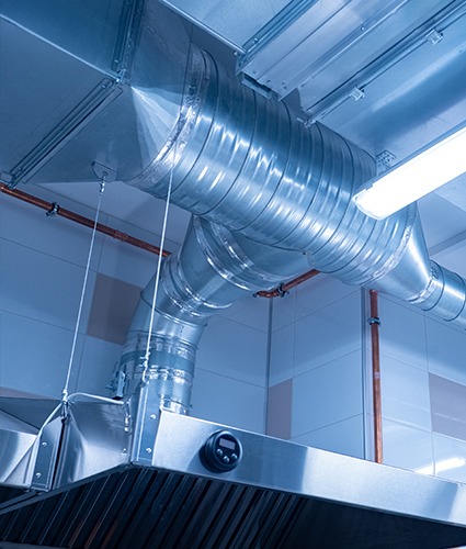 Bright metal ductwork and piping running above a commercial kitchen